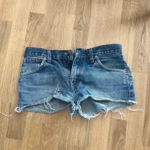 Levi's blå jeansshorts med fransar - Snygga blå jeansshorts från Levi's med hög midja och råa, fransiga kanter. Klassisk femficksmodell med slitna detaljer och ikonisk läderpatch bak. Perfekta för sommardagar och festivalhäng. Styla med en oversized tee eller crop top för en chill look.