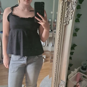 Svart linnetopp med knytband 157 - Supersöt svart linnetopp från 157 med smala axelband som knyts på axlarna och volangkant upptill. Toppen har en lätt rynkad struktur och luftig passform, perfekt för varma dagar. Enkel att matcha med jeans eller kjol för en chill look.