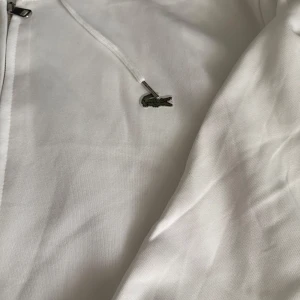 Vit hoodie från Lacoste med dragkedja - Säljer en fräsch vit hoodie från Lacoste med klassisk krokodillogga på bröstet. Tröjan har huva med snörning och dragkedja framtill. Materialet är mjuk bomull och passformen är normal. Perfekt för chill dagar eller när du vill ha en clean look.