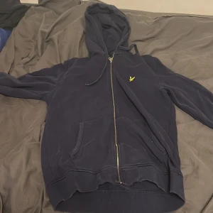 Mörkblå hoodie från Lyle & Scott - Säljer en mörkblå hoodie från Lyle & Scott med gul logga på bröstet. Tröjan har dragkedja, huva med snörning och två fickor framtill. Ribbad nederkant och ärmslut ger en schysst passform. storlek L men skulle säga liten i storlek 