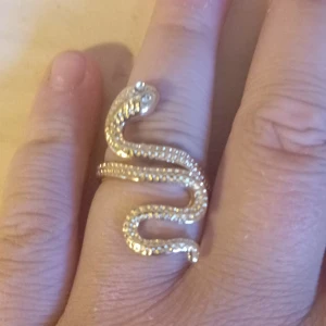 Guldig ormring i cool design - Unik ring i form av en slingrande orm i guldtonad metall. Ringen har detaljerad struktur längs kroppen och små stenar som ögon. Den är justerbar och ger en edgy vibe till din stil.