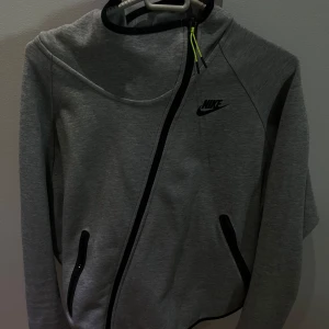 Grå hoodie från Nike med dragkedja - Snygg grå hoodie från Nike med svart logga på bröstet och cool diagonal dragkedja. Tröjan har huva, svarta detaljer vid fickorna och en gul snörning vid halsen. Perfekt för dig som gillar sportig och stilren look.