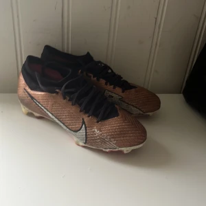 Nike Mercurial fotbollsskor elites - Snygga Nike Mercurial fotbollsskor i brons med svarta detaljer och svart snörning. Skorna har en låg profil och är tillverkade i syntetmaterial med texturerad yta för bättre bollkontroll. Perfekta för dig som vill sticka ut på planen. Fast pris nypris 2700kr