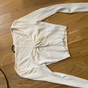 Vit ribbad långärmad topp med knyt - Säljer en vit ribbad långärmad topp med snygg v-ringning och knyt framtill. Toppen är croppad och har en smickrande passform som passar perfekt till jeans eller kjol. Materialet är mjukt och stretchigt för extra komfort.