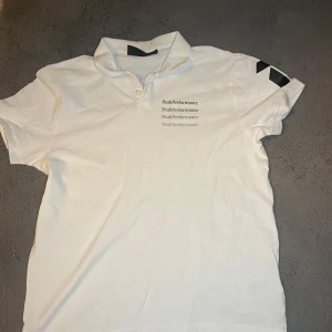 Vit Peak Performance t-shirt med krage - Vit t-shirt från Peak Performance i storlek S med klassisk krage och knappslå. Tryck med Peak Performance-loggan i flera rader på bröstet och svart detalj på ena ärmen. Skön bomullskvalitet och stilren look som funkar till jeans eller shorts.