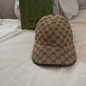 Gucci keps med GG-mönster - Snygg keps från Gucci i beige och brunt med klassiskt GG-monogram över hela kepsen. Justerbar rem bak i mörkbrunt läder och ikoniska gröna och röda ränder på sidan. Tillverkad i bomullscanvas med exklusiv känsla.         Pris kan diskuteras!