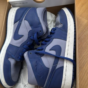 Nike Air Jordan 1 High blå/lila - Säljer ett par Nike Air Jordan 1 High sneakers i blått och ljuslila skinn. Klassisk siluett med högt skaft, perforerad tå och blå snörning. Snygg kontrast mellan de två färgerna och ikoniska Jordan-loggan på sidan. Perfekta för dig som gillar streetstyle.