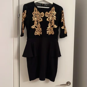 Svart kort klänning med guldpaljetter - Snygg svart kort klänning med guldiga paljettdetaljer framtill och på ärmarna. Klänningen har peplumvolanger vid midjan, halvärmar och en synlig dragkedja baktill. Perfekt för dig som vill sticka ut med glittriga detaljer och elegant siluett.