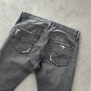 Vintage Levi's 501  - Grå vintage Levi's 501 jeans med slitna detaljer på bakfickorna och råa, fransiga benslut. Klassisk femficksmodell med rak passform och vintage vibe. Perfekta för dig som gillar en avslappnad och edgy stil.