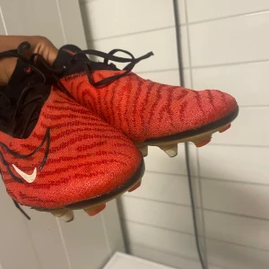 Nike Phantom GT2 röda fotbollsskor - Nike Phantom GT2 fotbollsskor i rött och svart med snyggt mönster och stora vita Swoosh-loggor på sidan och hälen. Skorna har snörning och en låg profil, perfekt för dig som vill ha grym touch och kontroll på planen. Ovandelen är i syntetmaterial och sulan har dobbar för gräs.