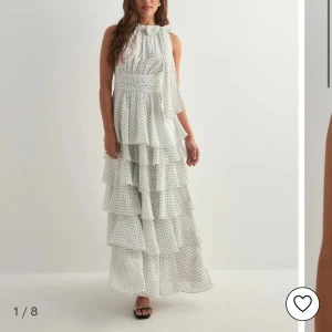 Vit långklänning med volanger och prickar - Supersöt vit långklänning med svarta prickar och flera lager volanger. Klänningen har hög hals med knyt och är ärmlös, vilket ger en fräsch och somrig vibe. Perfekt för dig som gillar romantisk stil och vill sticka ut med ett lekfullt mönster. Använd en gång på en middag men inga tecken på användning. Säljer då den är för liten. 💞