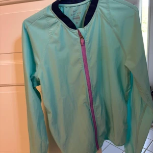 Nike Dri-Fit turkos vindjacka S - Nike Dri-Fit vindjacka i fräsch turkos färg med lila dragkedja och mörkblå krage. Jackan är lätt och har lång ärm, perfekt för aktiva dagar. Snygg sportig look och tillverkad i polyester som andas.