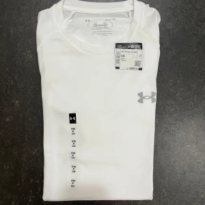 Vit t-shirt från Under Armour - Snygg vit t-shirt från Under Armour med klassisk rund hals och diskret logga på bröstet. Tillverkad i ett lätt och funktionellt syntetmaterial som andas, perfekt för träning eller en aktiv vardag. Modellen har korta ärmar och en loose passform.