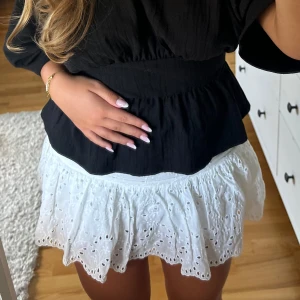 Vit kort kjol med broderade detaljer - Supersöt vit kort kjol med broderade hålmönster och volangkant. Kjolen har en luftig och lätt känsla, perfekt för varma dagar. Den sitter normalt i midjan och har en feminin, ungdomlig vibe med sina fina broderier och volanger.