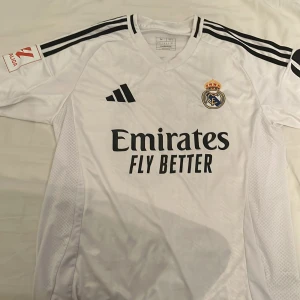 Real Madrid Mbappé 10 vit matchtröja - Snygg Real Madrid fotbollströja från Adidas i slim fit, storlek M. Vit med svarta detaljer och klassiska ränder på axlarna. Tryck med Mbappé och nummer 10 på ryggen, samt klubbmärke och sponsor på bröstet. Tillverkad i lätt och ventilerande polyester.