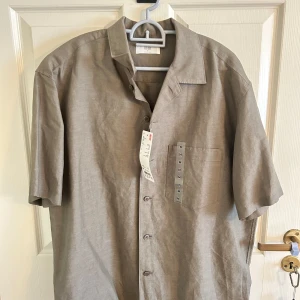 Olivgrön linneskjorta från Uniqlo  - Snygg olivgrön skjorta från Uniqlo i storlek M. Skjortan har korta ärmar, klassisk krage, bröstficka och knäppning framtill. Tillverkad i en skön mix av linne och bomull som ger en luftig känsla. Perfekt för varma dagar. Köptes för 399kr