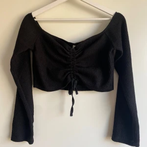 Svart croppad ribbad topp - Svart långärmad croppad topp med ribbad struktur och snörning framtill. Toppen har en bred, något offshoulder urringning och är perfekt för att skapa en trendig look. Snygg att styla med höga jeans eller kjol.