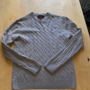 Merino kabelstickad tröja - Kabelstickad grå tröja från Morris i 100% merinoull. Klassisk rund halsringning och långärmad modell med blå broderad logga på bröstet. Perfekt för dig som gillar stilren och tidlös design med en touch av preppy. Säljer pga att den blivit för liten