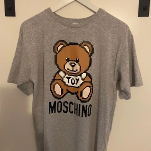 Grå Moschino t-shirt med nalletryck - Snygg grå t-shirt från Moschino med ett stort pixelinspirerat tryck av den ikoniska Moschino-nallen och texten MOSCHINO på framsidan. Klassisk rund hals och korta ärmar. Perfekt för dig som gillar streetstyle och coola prints. Tröjan är inhandlad från kidsbrandstore och är i gott skick! Storleken är 164 men den är rätt stor i sig passar mig som är 170 ish