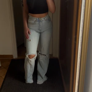 Ljusblå ripped high waist jeans - Säljer ett par ljusblå jeans med hög midja och raka ben. Jeansen har slitna detaljer och stora hål vid knäna för en cool, avslappnad vibe. Perfekta för dig som gillar en trendig och bekväm stil.