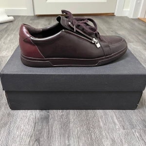 Mörkbruna/vinröda sneakers från Tiger of Sweden - Snygga mörkbruna/vinröda sneakers från Tiger of Sweden med stilren design och silverfärgad dragkedja på sidan. Skorna har snörning, rund tå och är tillverkade i skinn med en platt sula. Perfekta för dig som gillar minimalistisk/modern stil med en twist. Använda 1 gång. Nypris 2299. Originalbox tillkommer. 