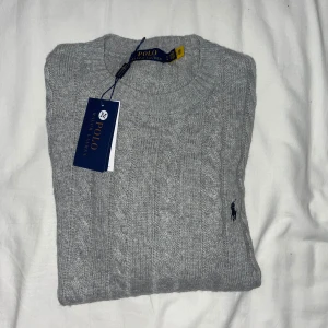Grå stickad tröja från Polo Ralph Lauren - Snygg grå kabelstickad tröja från Polo Ralph Lauren med klassisk rund halsringning och broderad logga på bröstet. Perfekt för lager-på-lager och chill stil. Mjuk och skön känsla, passar till allt.