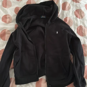 Svart hoodie från Polo Ralph Lauren - Svart hoodie från Polo Ralph Lauren med dragkedja och huva. Insidan är fodrad med mjuk fleece och tröjan har två fickor framtill samt den klassiska broderade loggan på bröstet. Perfekt för kyliga dagar och riktigt skön att ha på sig.