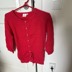 Röd kofta med trekvartsärmar - Röd kofta i stickad bomull. Koftan har trekvartsärmar, knappar framtill och en knytning i midjan. 