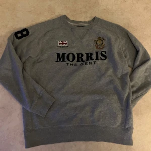 Grå sweatshirt från Morris The Gent - Snygg grå sweatshirt från Morris The Gent med tryck på bröstet, broderad patch och brittisk flagga. Tröjan har långa ärmar, ribbade muddar och klassisk passform. Perfekt för en stilren och avslappnad look.