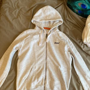 Superdry zip hoodie  - Snygg ljusgrå hoodie från Superdry Orange Label med dragkedja, huva med snörning och broderad logga på bröstet. Mjuk insida och två fickor framtill. Perfekt för chill dagar eller när du vill ha en clean streetstyle.