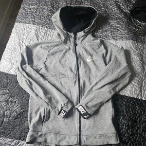 Grå Nike hoodie med dragkedja - Snygg grå hoodie från Nike med hel dragkedja och huva. Tröjan har långa ärmar och svarta muddar med vit Nike-logga. Perfekt för chill eller träning, med två fickor framtill och sportig look.