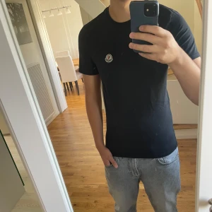 Moncler T-shirt svart - Svart t-shirt från Moncler med klassisk passform och rund hals. T-shirten har ett litet Moncler-märke broderat på bröstet och är tillverkad i mjuk bomull. Perfekt basic-plagg med stilren look och korta ärmar.