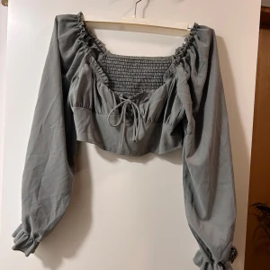 Grå croppad blus med knyt - Supersöt grå croppad blus med långa puffärmar och smockad överdel. Toppen har volangkant vid halsringningen och en snygg knytning framtill. Perfekt för dig som gillar romantisk och trendig stil.