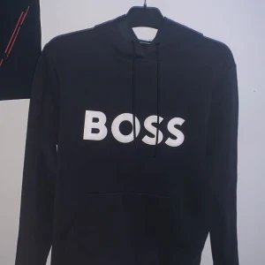 Svart hoodie från BOSS med tryck - Svart hoodie från BOSS med stor vit BOSS-logga på bröstet. Tröjan har huva med snörning, lång ärm och en klassisk känguruficka framtill. Materialet är mjuk bomull och polyester, perfekt för chill dagar. Snygg och enkel design som ger en clean look.tar emot byten finns i Norrköping kan gå ner snabb affär