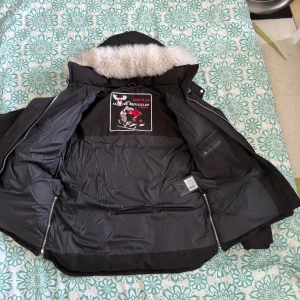 Svart dunjacka Moose Knuckles med pälskrage - Säljer en svart dunjacka från Moose Knuckles med snygg pälskantad huva. Jackan har flera dragkedjor, logga på ärmen och innerficka med dragkedja. Perfekt för kalla dagar och har en riktigt schysst streetstil. Fodrad och med detaljer i silver.