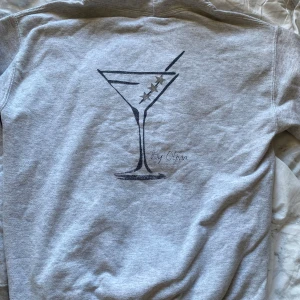 Grå hoodie med tryck på ryggen  - Grå hoodie med ett svart tryck av ett martiniglas på ryggen. Tröjan har huva, magficka och långa ärmar🪩💖 originalpris 760kr
