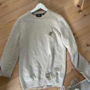 Beige stickad tröja från Lyle & Scott - Säljer en stilren beige fin stickad tröja från Lyle & Scott med deras ikoniska logga på bröstet. Tröjan har långa ärmar och ribbade muddar vid ärmslut och nederkant. använd en gång så näst intill helt ny