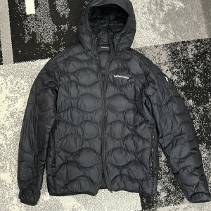 Peak Performance jacka  - Säljer en svart dunjacka från Peak Performance med dragkedja och huva. Jackan har ett quiltat mönster och är perfekt för hösten/våren. Den har långa ärmar och en stilren design med logotyp på bröstet och ärmen.