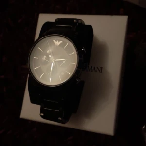 Svart klocka från Emporio Armani - Snygg svart klocka från Emporio Armani med stilren design. Klockan har en svart urtavla med gulddetaljer och flera små urtavlor för extra funktionalitet. Perfekt accessoar för den stilmedvetna. man måste bara ställa in klockan i en butik.