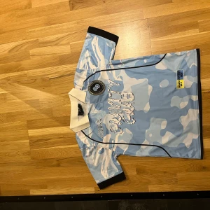 Ljusblå kamouflage fotbollströja - Säljer en ljusblå kamouflage fotbollströja med vit krage och korta ärmar. Tröjan har svarta detaljer och texten 'Believe in County Club' på baksidan. Nummer 33 är tryckt stort på ryggen. Perfekt för fotbollsfans som vill sticka ut!