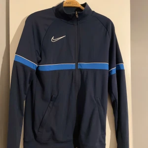 Mörkblå träningsjacka från Nike - Säljer en mörkblå zip tröja från Nike med ljusblåa detaljer. Två stycken fickor med dragkedjor 