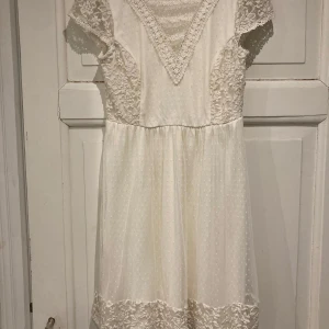 Vit examensklänning från River Island - Charmig vit klänning från River Island med vackra spetsdetaljer och korta ärmar. Klänningen har en v-ringad design med broderade blommor och ett prickigt mönster. Perfekt för en somrig look!