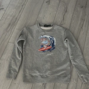 Grå sweatshirt från Polo Ralph Lauren - Säljer en grå sweatshirt från Polo Ralph Lauren med ett tryck av en surfande björn på framsidan. Tröjan har långa ärmar och rund halsringning. Perfekt för en avslappnad stil.