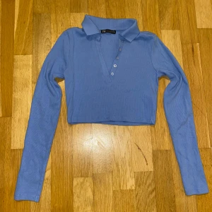 Blå croppad polotopp från Zara - Säljer en snygg blå croppad polotopp från Zara. Toppen har långa ärmar och knappar framtill. Perfekt för en stilren look.