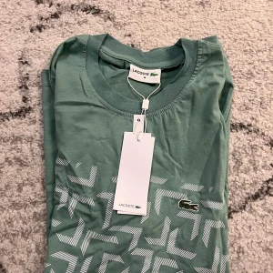 Grön t-shirt från Lacoste - Snygg grön t-shirt från Lacoste med ett unikt geometriskt mönster i vitt. T-shirten har en klassisk rund hals och korta ärmar. Perfekt för en stilren och avslappnad look.