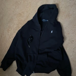 Svart hoodie från Polo Ralph Lauren - Säljer en stilren svart hoodie från Polo Ralph Lauren med dragkedja och den klassiska loggan broderad i vitt på bröstet. Hoodien har en bekväm passform och är tillverkad i mjukt material. Passar även perfekt på storlek L.