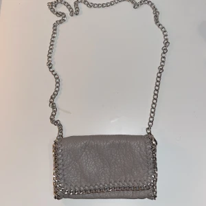 Grå axelväska med kedjedetaljer - Snygg grå crossbody-väska med kedjedetaljer och magnetlås. Väskan har en kedjerem och en dragkedja inuti för extra säkerhet. 