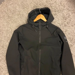 Svart windbreaker med huva - Snygg svart vindjacka med huva och dragkedja framtill. Jackan har en stilren design med två fickor och justerbara snören vid huvan. Perfekt för blåsiga dagar.