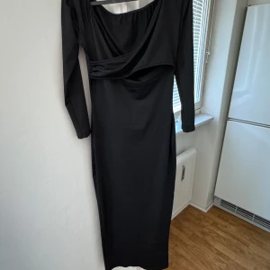 Svart långklänning med drapering - Elegant svart långklänning med draperad detalj framtill. Klänningen har långa ärmar och en stilren design som passar perfekt för festliga tillfällen.