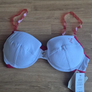 Ny BH vit och röd bh med Snobben 80B - Säljer en söt vit bh med röda detaljer och ett tryck med Snobben / Snoopy på. Bh:n har justerbara axelband med mönster och en dekorativ detalj mellan kuporna. Perfekt för den som vill ha något unikt och lekfullt. Dessa underkläder var The Shit i början av 2000-talet och gör nu en revival igen. Storlek 80B. Är helt ny, kommer från konkurslager och har orginaletiketten kvar. För dyrt? Kom med bud.